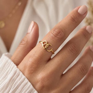 Op de afbeelding: Een goudkleurige ring met twee in elkaar grijpende letters, B en C, die het centrale ontwerpelement vormen. De band is dun en rond, met een gepolijste metalen afwerking die de vinger omsluit met een eenvoudige en strakke uitstraling.