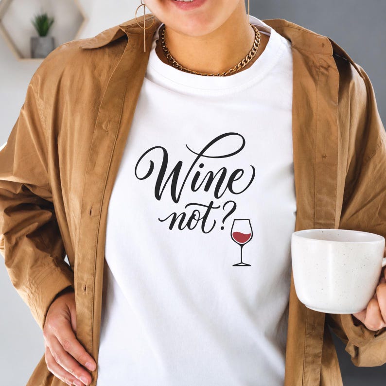 May include: A white t-shirt featuring the black cursive text "Wine not?" and a small graphic of a red wine glass. The shirt is worn under a tan button-down shirt, paired with a gold chain necklace and gold hoop earrings for a complete look.