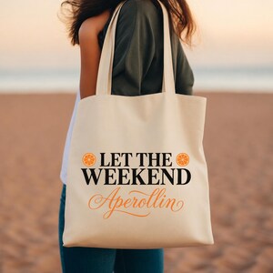 May include: A cream-colored canvas tote bag featuring the black text LET THE WEEKEND and the orange cursive text Aperollin, flanked by two orange citrus slice icons. The bag has two long shoulder straps and a rectangular body shape.