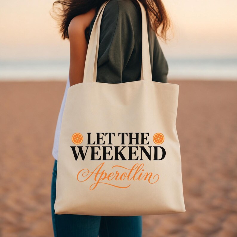 May include: A cream-colored canvas tote bag featuring the black text LET THE WEEKEND and the orange cursive text Aperollin, flanked by two orange citrus slice icons. The bag has two long shoulder straps and a rectangular body shape.