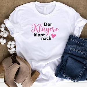 May include: A white short-sleeved t-shirt featuring the black and pink text 'Der Kl&uuml;gere kippt nach' with a small pink heart. The shirt is displayed alongside dark blue denim jeans and tan suede ankle boots with a block heel.