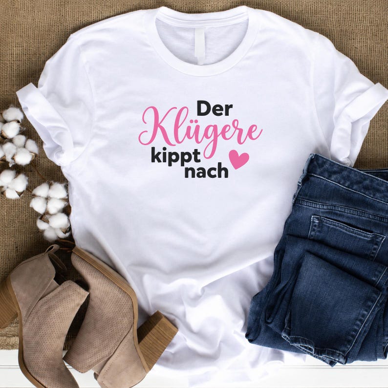 May include: A white short-sleeved t-shirt featuring the black and pink text 'Der Kl&uuml;gere kippt nach' with a small pink heart. The shirt is displayed alongside dark blue denim jeans and tan suede ankle boots with a block heel.