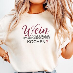 May include: A cream-colored t-shirt featuring dark maroon and black text that reads Wein KALT STELLEN IST AUCH IRGENDWIE KOCHEN with a small heart icon and a wine glass graphic. The shirt has a crew neckline and short sleeves, worn with gold bracelets.