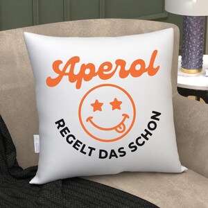 May include: A square white throw pillow featuring the orange text Aperol at the top, a central orange smiley face with star-shaped eyes and a tongue sticking out, and the black text REGELT DAS SCHON curved underneath the smiley face.