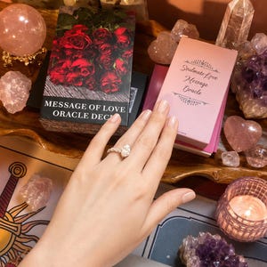 Puede incluir: Una caja de mazo de cartas Message of Love Oracle con rosas rojas y una caja de Soulmate Love Message Oracle en rosa claro. Un anillo de plata con una piedra en forma de pera se lleva en un dedo. Los objetos est&aacute;n rodeados de cristales y una vela.