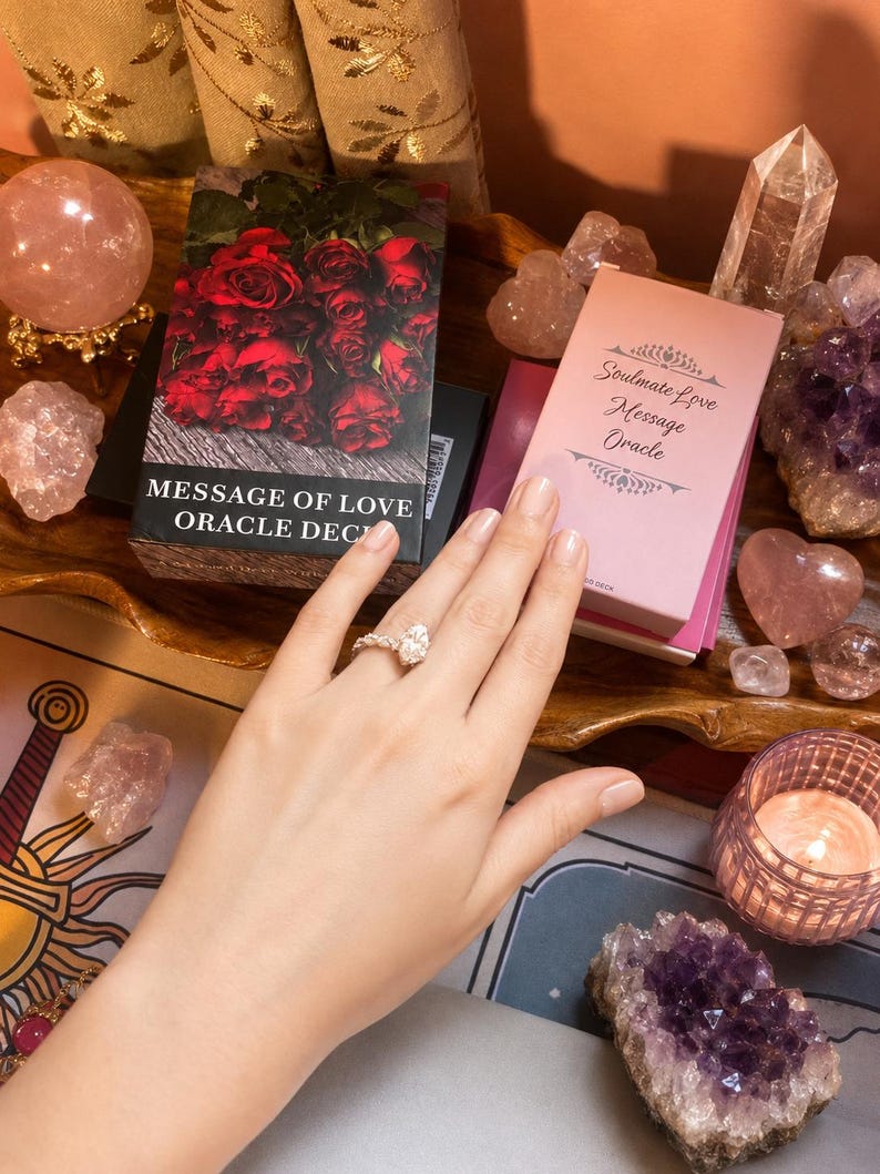 Puede incluir: Una caja de mazo de cartas Message of Love Oracle con rosas rojas y una caja de Soulmate Love Message Oracle en rosa claro. Un anillo de plata con una piedra en forma de pera se lleva en un dedo. Los objetos est&aacute;n rodeados de cristales y una vela.