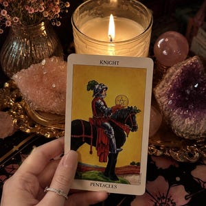 Puede incluir: Una carta de tarot titulada Knight of Pentacles que presenta la ilustraci&oacute;n de un caballero con armadura plateada montando un caballo negro mientras sostiene una moneda de oro con un s&iacute;mbolo de pentagrama. La carta tiene un fondo amarillo.