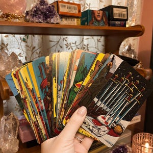 Puede incluir: Un abanico de cartas de tarot sostenido en una mano, con la carta del Diez de Espadas destacada en primer plano. La baraja muestra varias ilustraciones coloridas, incluyendo figuras, paisajes e im&aacute;genes simb&oacute;licas en cada carta.