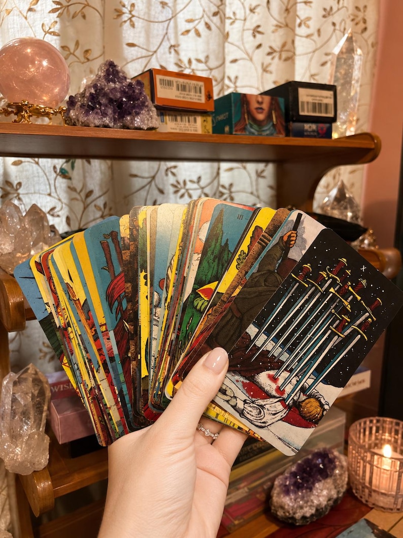 Puede incluir: Un abanico de cartas de tarot sostenido en una mano, con la carta del Diez de Espadas destacada en primer plano. La baraja muestra varias ilustraciones coloridas, incluyendo figuras, paisajes e im&aacute;genes simb&oacute;licas en cada carta.