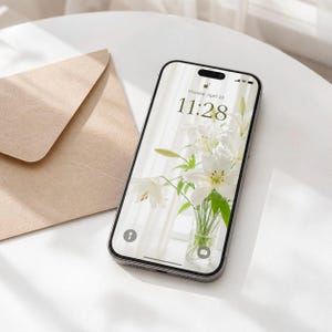 May include: A smartphone with a black frame displaying a lock screen with the time 11:28 and the date Monday, April 28. The screen background features a clear glass vase filled with white lilies. A tan paper envelope sits next to the smartphone.