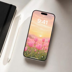 May include: A smartphone with a black frame displaying a lock screen with a sunset over a field of pink tulips, showing the time 9:41 and the date Monday, June 10. A white stylus and a black notebook are positioned to the left of the device.