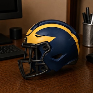 May include: A navy blue football helmet featuring a prominent yellow wing design on the sides and a dark gray face mask. The helmet has a matte finish and includes a small yellow letter M logo on the front center above the face mask.