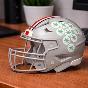 May include: A silver football helmet featuring a gray face mask and a central red, white, and black stripe. The side of the helmet is decorated with a cluster of circular white stickers, each displaying a small green leaf graphic.