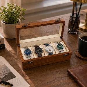 Puede incluir: Una caja expositora de madera rectangular con tapa de cristal transparente y forro interior de color crema. La caja cuenta con cuatro compartimentos individuales, cada uno con un reloj de pulsera con un color de esfera único, incluyendo azul, negro, blanco y verde.