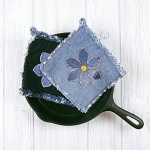 Puede incluir: Dos agarraderas de olla de mezclilla azul con un diseño de flor, una con un centro amarillo, sobre una sartén de hierro fundido negra.