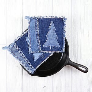 Puede incluir: Dos agarraderas de olla de mezclilla azul con borde deshilachado y un diseño de árbol bordado en blanco. Las agarraderas están sobre una sartén de hierro fundido negra.
