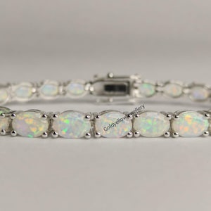 Könnte beinhalten: Ein silberfarbenes Tennisarmband mit einer Reihe ovaler, irisierender weißer Opale, die in Metallfassungen eingefasst sind. Das Armband verfügt über einen sicheren Kastenschloss-Verschluss und zeigt den Text Goldyellowjewellery auf den mittleren Gliedern.