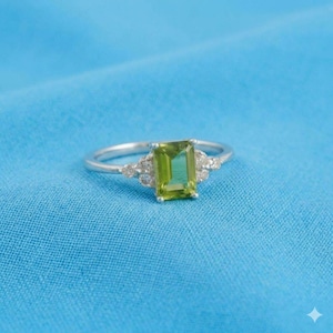 May include: A silver ring featuring a rectangular, emerald-cut lime green gemstone at the center. The band is accented with three small, round, clear stones on each side of the central setting, creating a symmetrical design with a polished metal finish.