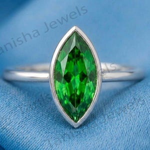 Puede incluir: Un anillo de plata con una piedra preciosa de color verde vibrante en talla marquesa, montada en un engaste de bisel liso. La banda es de plata pulida y el texto "anisha Jewels" es visible al fondo. El diseño resalta la forma alargada de la piedra.