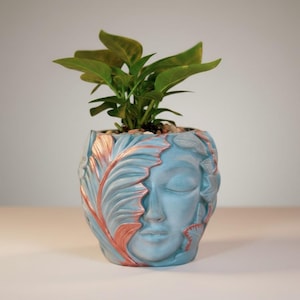 May include: A light blue planter shaped like a human face with closed eyes, adorned with raised leaf patterns highlighted in metallic copper. A green leafy plant grows from the top of the vessel, which features a smooth, matte finish.