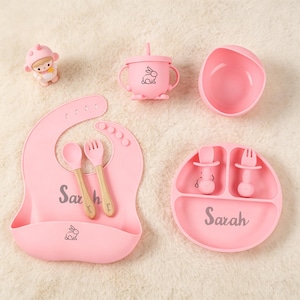 May include: A pink baby feeding set featuring a bib, bowl, plate, cup, spoon, and fork. The set includes a bib with the name "Sarah" and a bunny graphic. The cup and plate also have bunny graphics. The set is made of silicone.