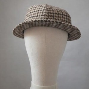 May include: A brown and black houndstooth pattern hat with a short brim. The hat is displayed on a mannequin head. The hat's classic design is suitable for various occasions.
