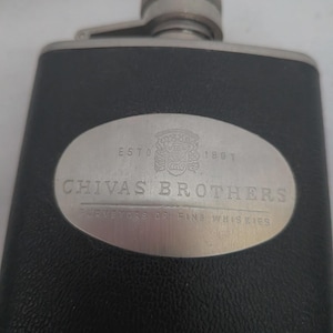 May include: A black leather-wrapped stainless steel flask featuring a central oval metal plate engraved with the Chivas Brothers logo, the text ESTD 1801, and the words PURVEYORS OF FINE WHISKIES. The flask has a screw-top cap at the top.