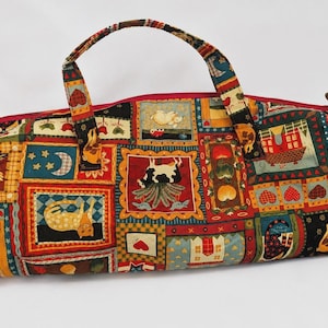 May include: A cylindrical fabric duffel bag featuring a colourful patchwork pattern with various illustrations including a cat, a cow, hearts, stars, and moons. The bag has two fabric handles and a red zip closure along the top edge.