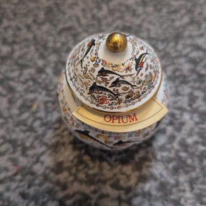 Può includere: Un barattolo in ceramica bianca con coperchio abbinato, caratterizzato da un pomello dorato e un motivo colorato di delfini e vita marina. Un'etichetta beige curva con il testo rosso OPIUM è fissata sulla parte anteriore del barattolo rotondo.