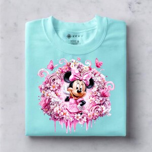 Puede incluir: Una camiseta de color turquesa claro con un gr&aacute;fico central de Minnie Mouse con un lazo y vestido de lunares rosas, rodeada por una densa disposici&oacute;n circular de flores rosas, peque&ntilde;as mariposas y elementos decorativos de l&iacute;neas curvas.