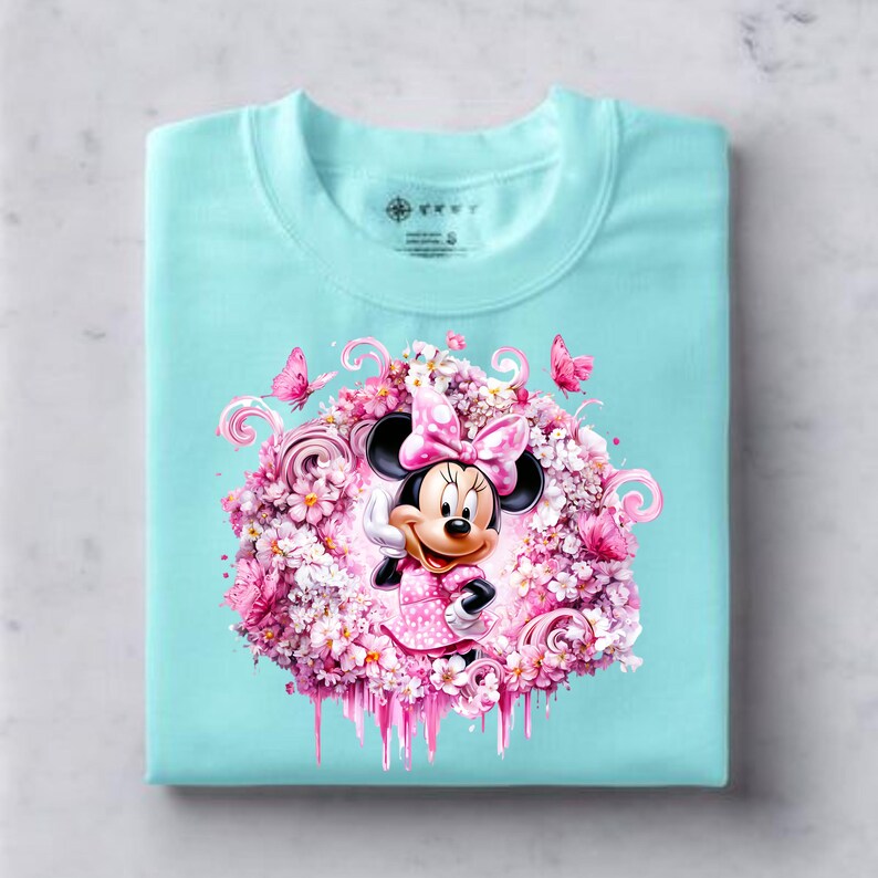 Puede incluir: Una camiseta de color turquesa claro con un gr&aacute;fico central de Minnie Mouse con un lazo y vestido de lunares rosas, rodeada por una densa disposici&oacute;n circular de flores rosas, peque&ntilde;as mariposas y elementos decorativos de l&iacute;neas curvas.