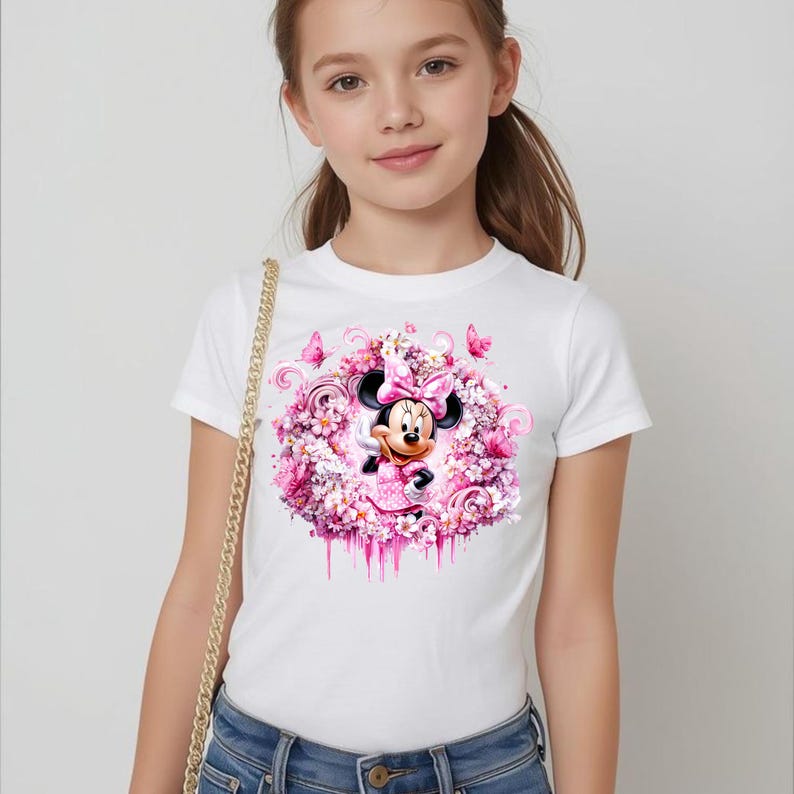 Puede incluir: Una camiseta blanca de manga corta con un gr&aacute;fico central de Minnie Mouse rodeada por un arreglo circular de flores rosas, mariposas y patrones en espiral. El dise&ntilde;o incluye un efecto de pintura que gotea en el borde inferior.