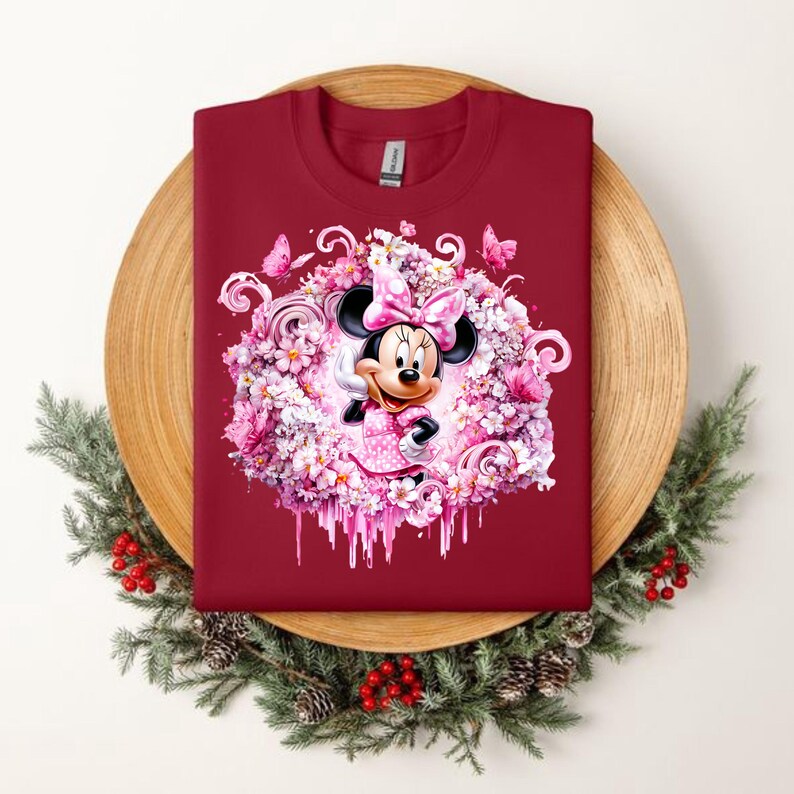 Puede incluir: Una camiseta de cuello redondo color rojo oscuro con un gr&aacute;fico central de Minnie Mouse rodeada por una corona de flores rosas, mariposas y patrones en espiral. El dise&ntilde;o incluye un efecto de pintura que gotea en la base de la corona.