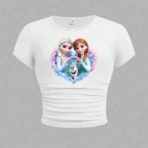 May include: White crop top featuring a colorful graphic of two animated female characters forming a heart shape with their hands, with a snowman in the center. The design is set against a floral background.