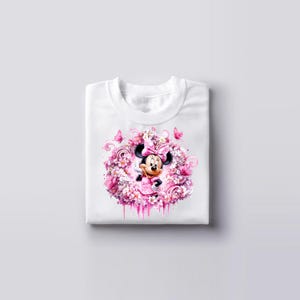 Puede incluir: Una camiseta blanca con un gr&aacute;fico central de Minnie Mouse rodeada por un denso grupo de flores rosas y blancas, peque&ntilde;as mariposas y patrones en espiral. El dise&ntilde;o est&aacute; representado en tonos rosa, blanco y magenta suave.