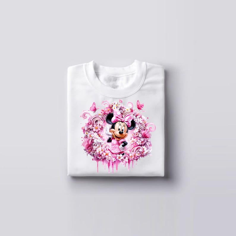 Puede incluir: Una camiseta blanca con un gr&aacute;fico central de Minnie Mouse rodeada por un denso grupo de flores rosas y blancas, peque&ntilde;as mariposas y patrones en espiral. El dise&ntilde;o est&aacute; representado en tonos rosa, blanco y magenta suave.