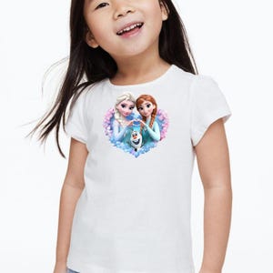 May include: White t-shirt featuring a heart-shaped graphic with two animated female characters and a snowman. The characters are in light blue dresses, with one having blonde hair and the other with red hair. The snowman is white with a carrot nose.