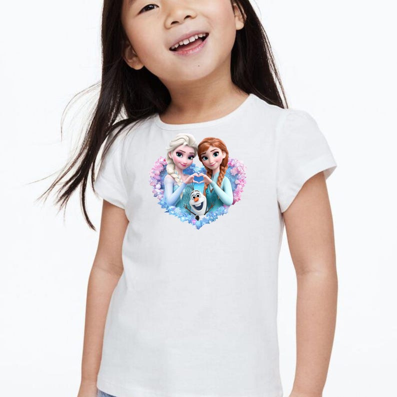 May include: White t-shirt featuring a heart-shaped graphic with two animated female characters and a snowman. The characters are in light blue dresses, with one having blonde hair and the other with red hair. The snowman is white with a carrot nose.