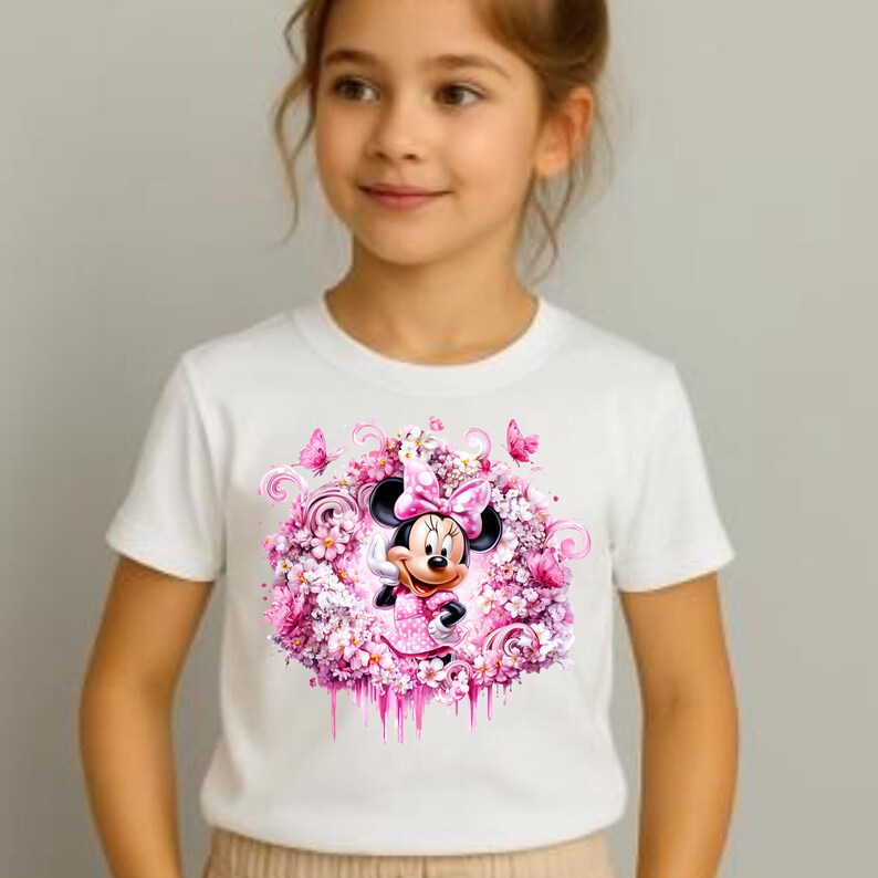 Puede incluir: Una camiseta blanca de manga corta con un estampado gr&aacute;fico central de Minnie Mouse rodeada por un vibrante arreglo de flores rosas, espirales y peque&ntilde;as mariposas. El dise&ntilde;o es circular con un efecto de pintura que gotea en la parte inferior.