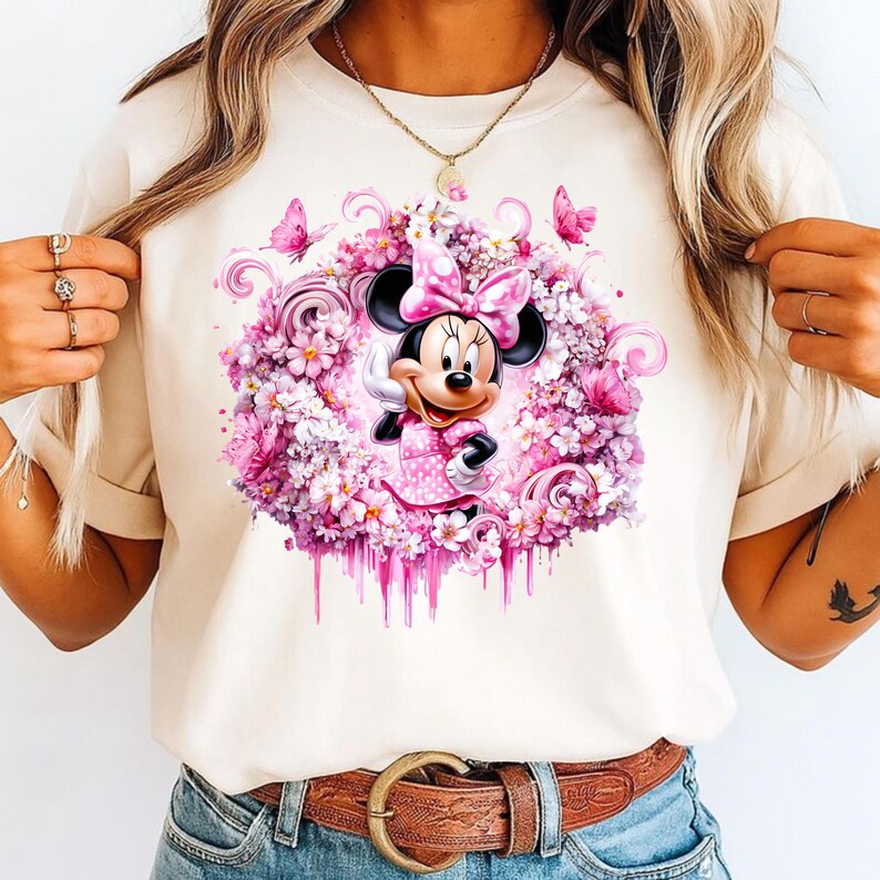 Puede incluir: Una camiseta de color crema con un gr&aacute;fico central de un rat&oacute;n de dibujos animados con un lazo de lunares rosa y un vestido, rodeado por una corona de flores rosas y moradas, patrones de remolinos y peque&ntilde;as mariposas con efecto goteo.