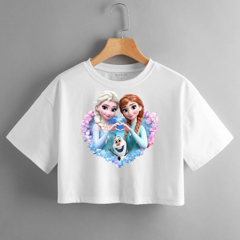 May include: White crop top featuring a colorful graphic of two animated princesses forming a heart shape with their hands, with a smiling snowman in the center. The shirt is hanging on a wooden hanger.