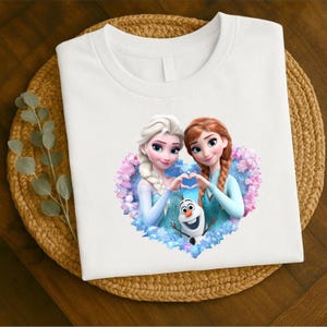 May include: White t-shirt featuring a heart-shaped graphic with two animated female characters and a snowman. The characters are in light blue dresses, forming a heart shape with their hands. The graphic is surrounded by pink and blue floral elements.