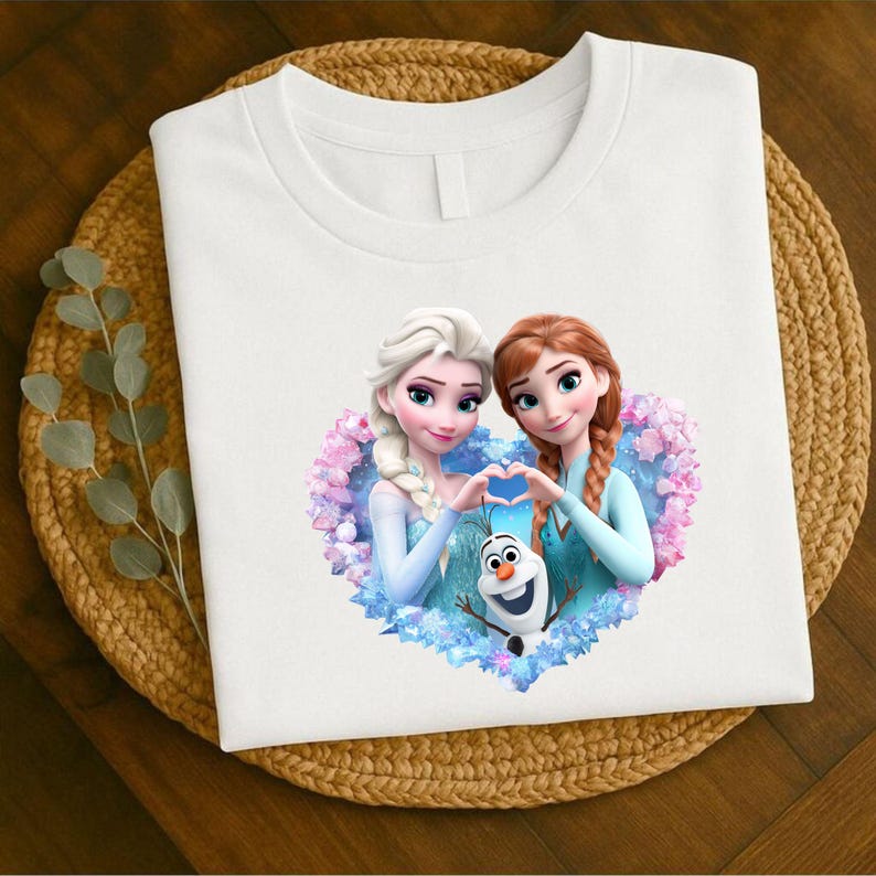 May include: White t-shirt featuring a heart-shaped graphic with two animated female characters and a snowman. The characters are in light blue dresses, forming a heart shape with their hands. The graphic is surrounded by pink and blue floral elements.