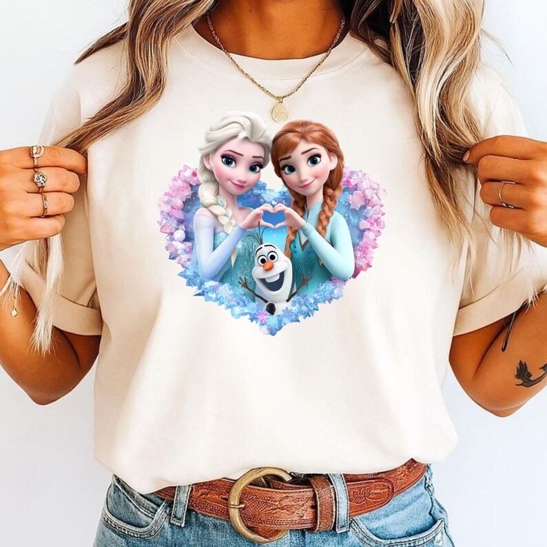 May include: Cream-colored t-shirt featuring a heart-shaped design with Elsa, Anna, and Olaf from the movie Frozen. The characters are in their signature outfits, surrounded by blue and purple floral accents. The sisters are making a heart shape with their hands.