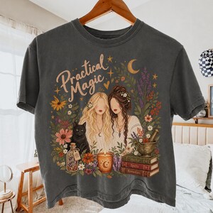 May include: A charcoal gray t-shirt featuring a graphic print with the text "Practical Magic" above two figures with long hair, a black cat, a candle, a mortar and pestle, and a stack of three books labeled "Herbs," "Spells," and "Remedies."