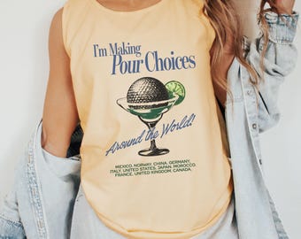 May include: A butter yellow sleeveless tank top featuring a graphic of a cocktail glass with a lime wedge and the text I'm Making Pour Choices Around the World, followed by a list of countries including Mexico, Norway, China, Germany, Italy, and others.