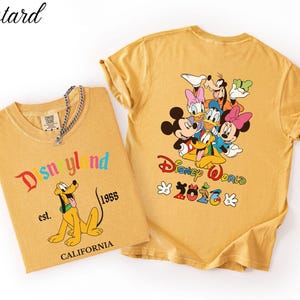 May include: Two mustard yellow t-shirts. The left shirt features Pluto, the text Disneyland, est. 1955, and CALIFORNIA. The right shirt displays a group of Disney characters, the text Disney World, and 2026 with Mickey-themed icons.