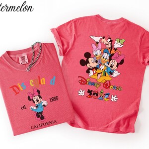 May include: Two watermelon-colored t-shirts. The left shirt features Minnie Mouse with the text Disneyland est. 1955 CALIFORNIA. The right shirt displays a group of Disney characters with the text Disney World 2026 and colorful accents.