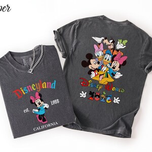 May include: Two charcoal gray t-shirts. The left shirt features Minnie Mouse, the text Disneyland, est. 1955, and CALIFORNIA. The right shirt displays a group of Disney characters, the text Disney World, and 2026 with Mickey-shaped icons.