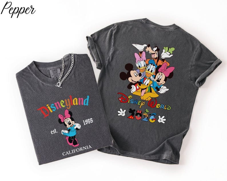 May include: Two charcoal gray t-shirts. The left shirt features Minnie Mouse, the text Disneyland, est. 1955, and CALIFORNIA. The right shirt displays a group of Disney characters, the text Disney World, and 2026 with Mickey-shaped icons.